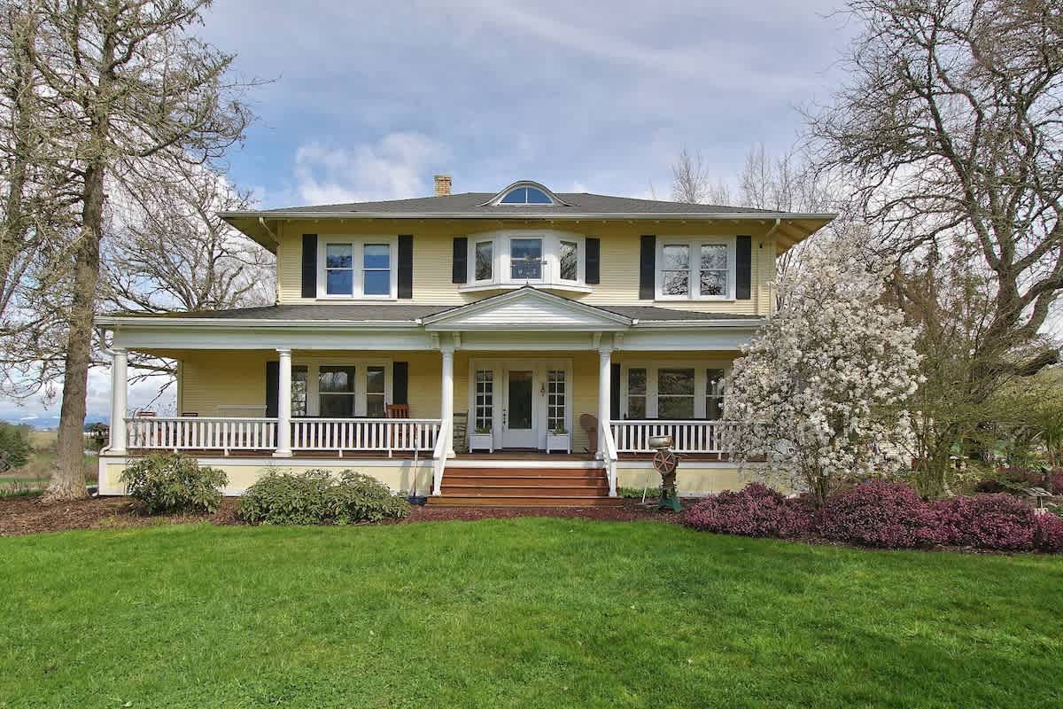 Grand yellow farm home with white trim, black shutters, and a wide wrap-around porch with white columns and railings. The house is surrounded by green grass, trees, and flowering bushes under a partly cloudy sky.