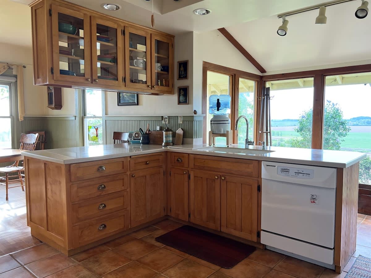 Spacious kitchen with L-shaped light wood cabinetry and light countertops, featuring a sink under large windows that provide ample natural light and views of the outdoors.