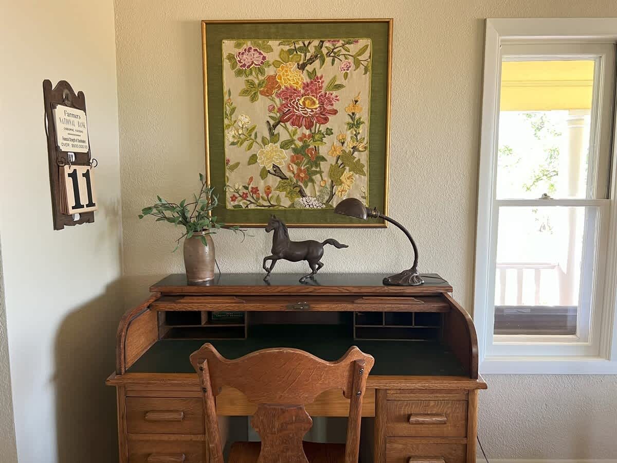 A close-up of a classic wooden roll-top desk with a green felt writing surface, adorned with a bronze horse figurine and a vintage desk lamp, positioned beneath a colorful framed floral artwork.