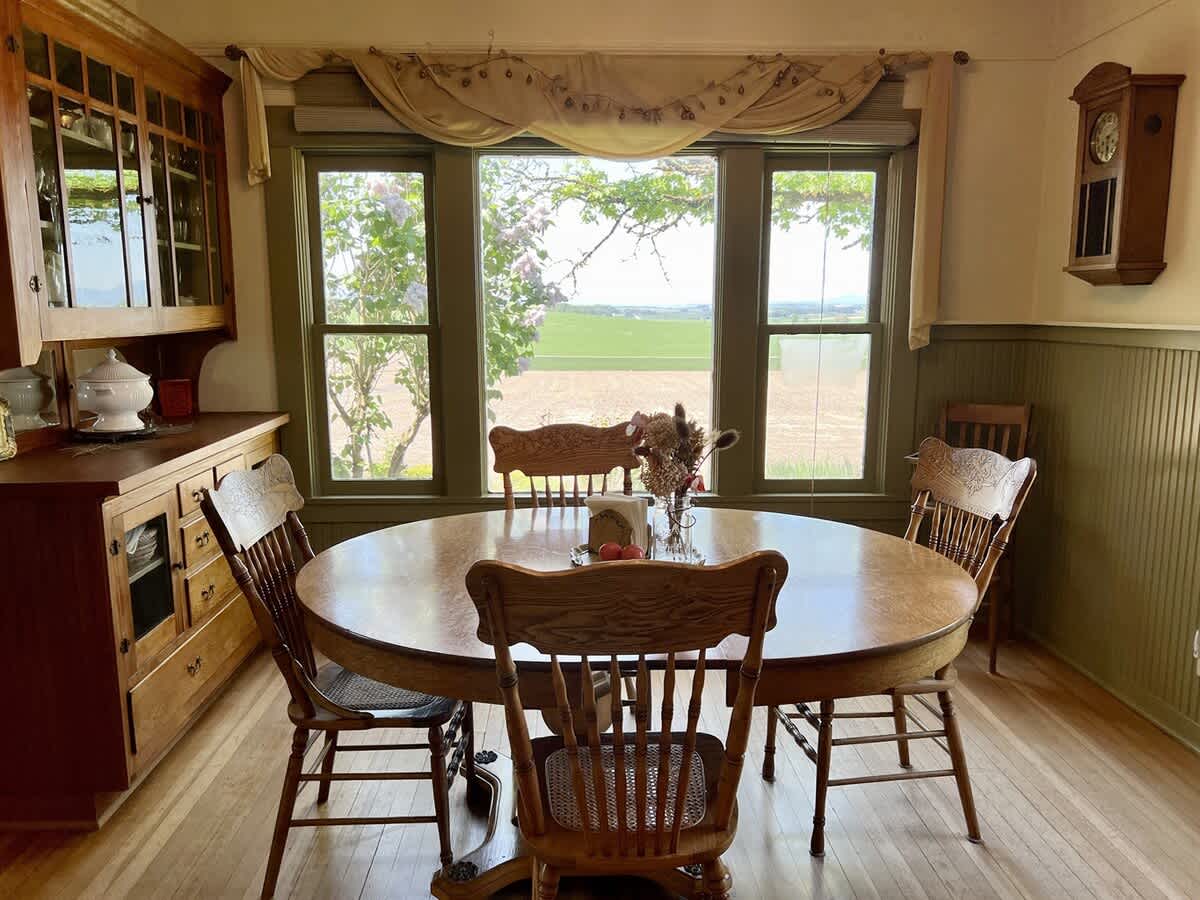 Bright dining room with a round wooden table and matching chairs, positioned in front of a wide window offering scenic views of the surrounding fields and trees.