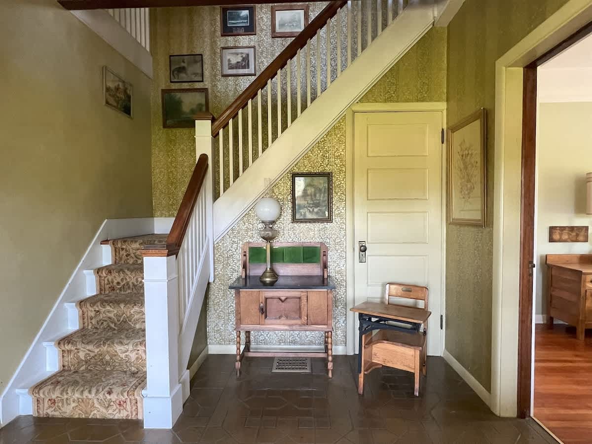 A classic foyer features a carpeted staircase with white balusters and a dark wooden handrail, complemented by patterned wallpaper and an antique wooden console table with a lamp.