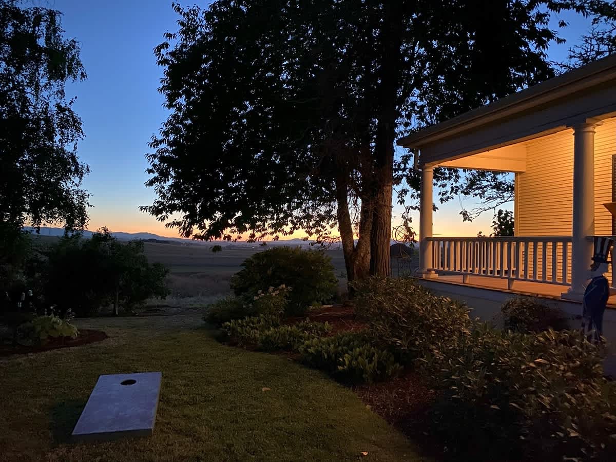 The property's expansive green lawn is illuminated by the soft glow of dusk, revealing a cornhole board in the foreground and the side of the house with a lit porch in the background, framed by large trees and a colorful horizon.