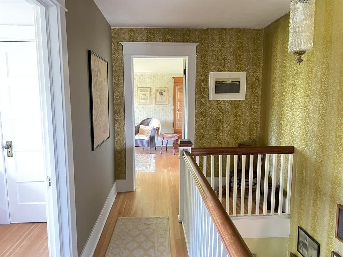Hallway or landing area with light wood floors and patterned wallpaper. A staircase railing is visible on the right, and a doorway leads to another room with a sitting area.