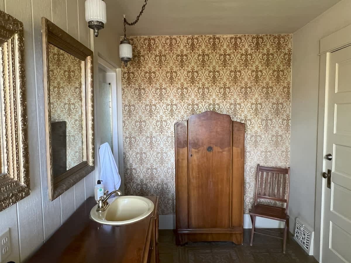 A vintage bathroom showcasing a dark wood vanity with a cream sink, ornate mirrors, a tall wooden armoire, and a wooden chair, all set against patterned wallpaper and floor tiles.