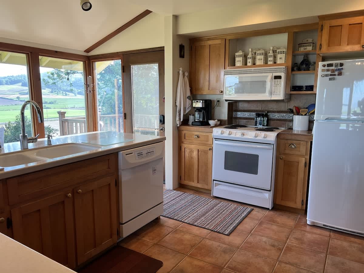Bright kitchen with light wood cabinets, white appliances including a stove, microwave, and refrigerator, and a large window overlooking a green landscape. A double sink is set into a light-colored countertop.