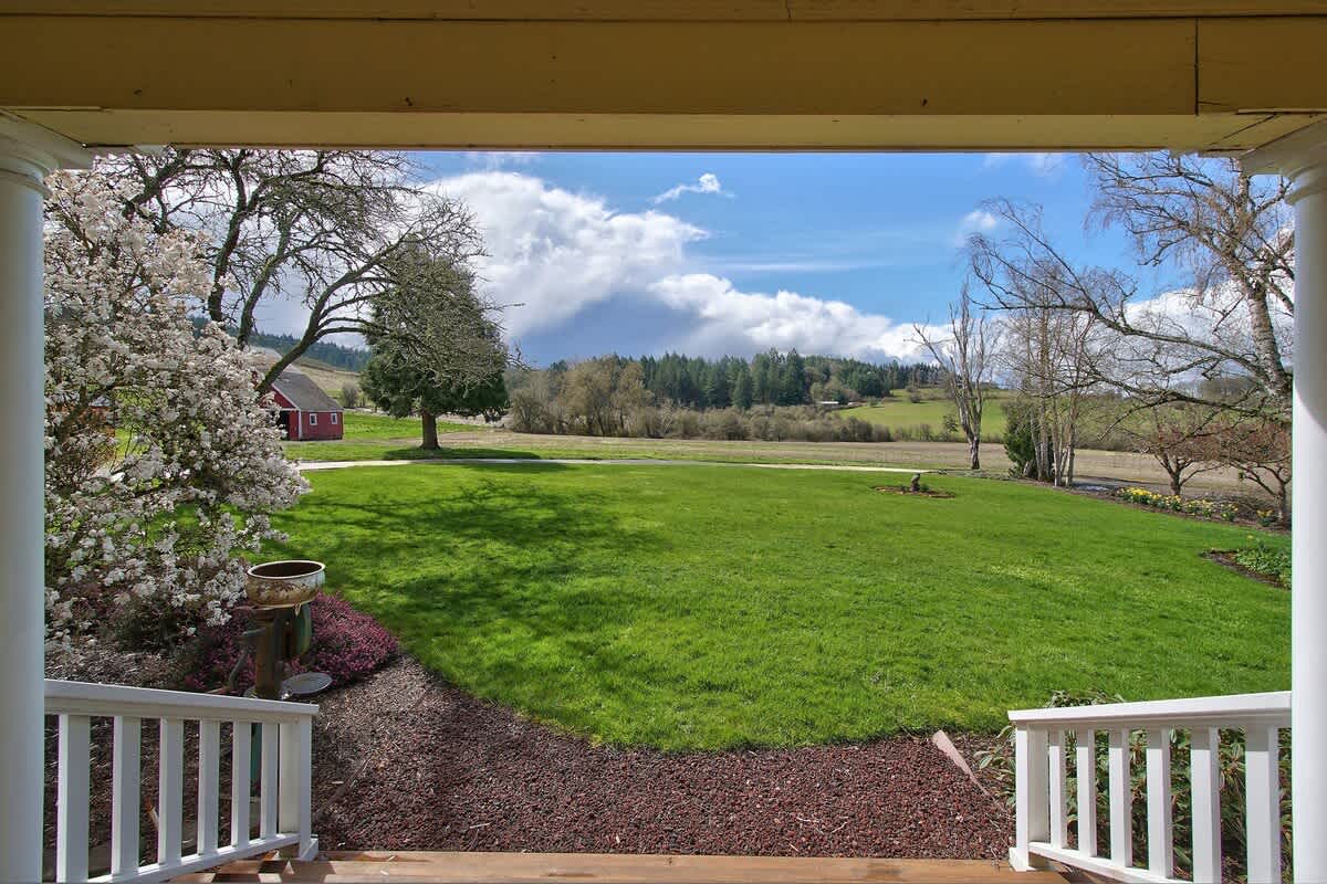 A wide-angle view from the porch captures a vast, vibrant green lawn stretching towards a red barn and scattered trees, all framed by a dramatic sky with large, fluffy white clouds.