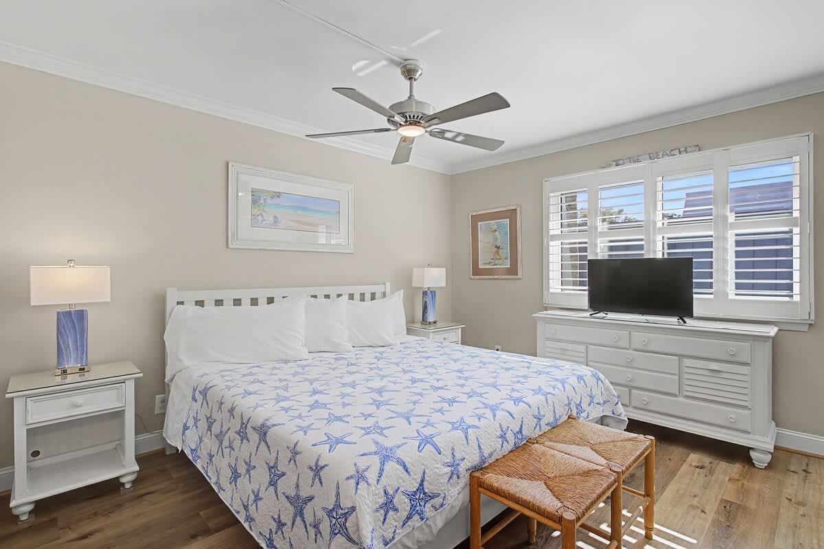 Relax in your coastal-inspired bedroom with soft starfish bedding, gentle ceiling fan, and natural light streaming through plantation shutters.