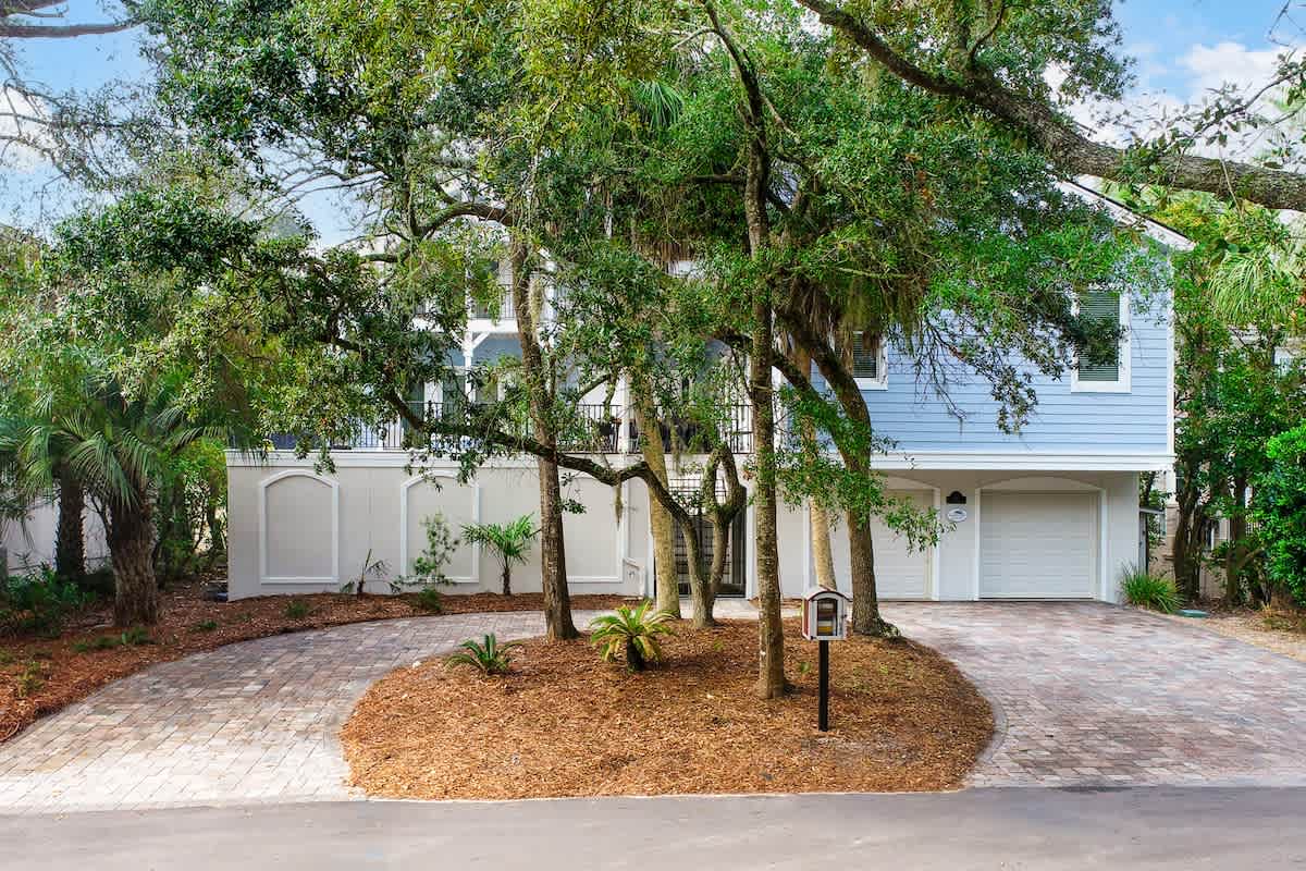 Modern white vacation home nestled among mature oak trees with circular brick driveway and tropical landscaping.