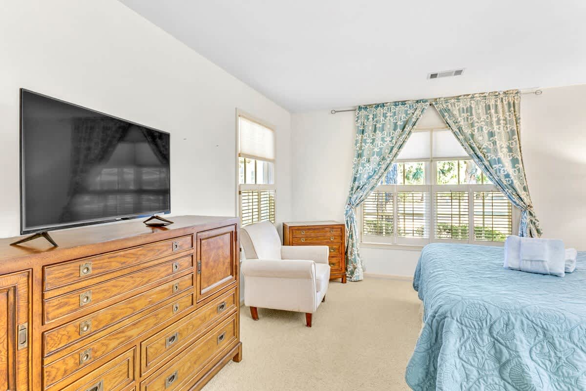 Unwind in your spacious bedroom with cozy blue bedding, large TV for relaxing evenings, and charming curtains framing peaceful garden views.
