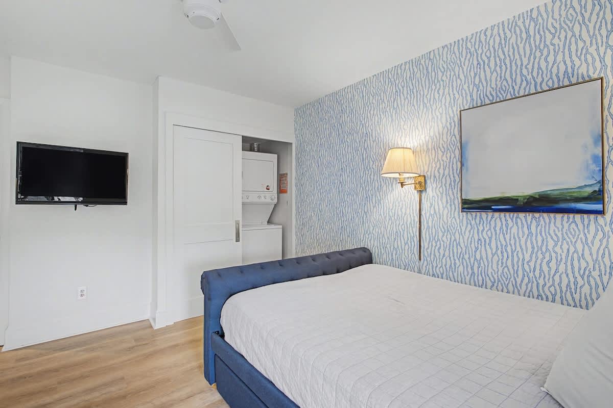 Relax in your serene bedroom with coastal-inspired decor, featuring a comfortable bed, wall-mounted TV, and convenient in-room laundry facilities.