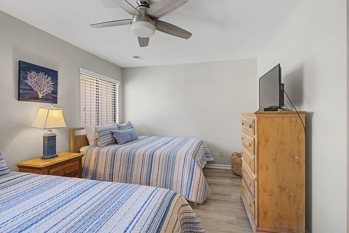 Relax in your peaceful twin bedroom with coastal decor, gentle ceiling fan, and warm natural light streaming through windows.