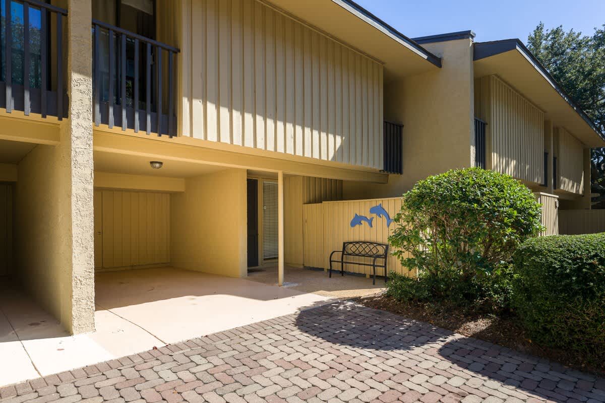 Modern vacation rental building with covered ground-floor patio and upstairs balcony, surrounded by manicured landscaping and brick pathways.