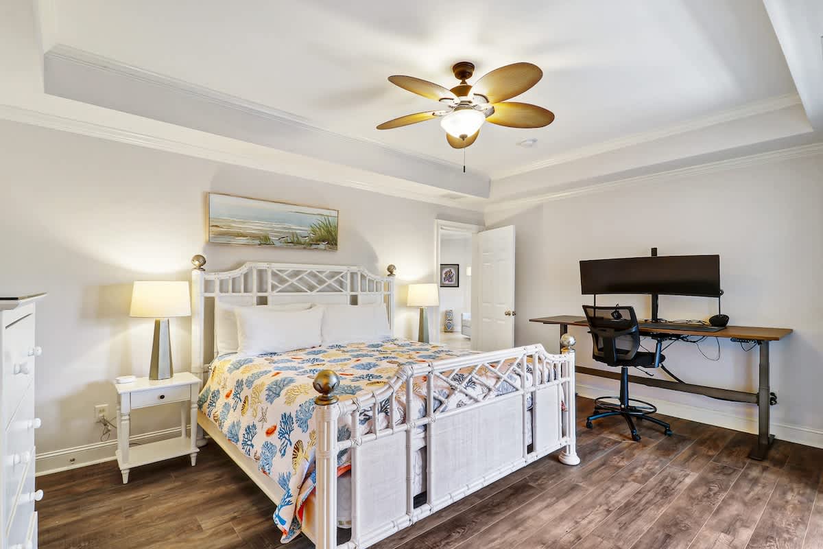 Your coastal-themed bedroom offers comfort with a stylish white bed, warm ceiling fan, and convenient workspace for productive stays.