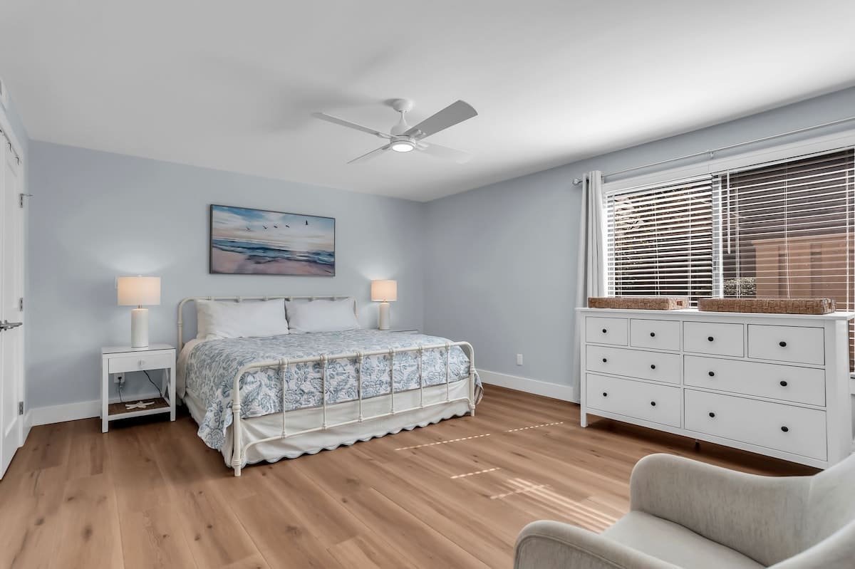 A serene primary bedroom retreat featuring a plush king bed dressed in soft coastal linens and soothing neutral tones. Light wood flooring and clean white furnishings create a bright, airy atmosphere perfect for restful nights.
