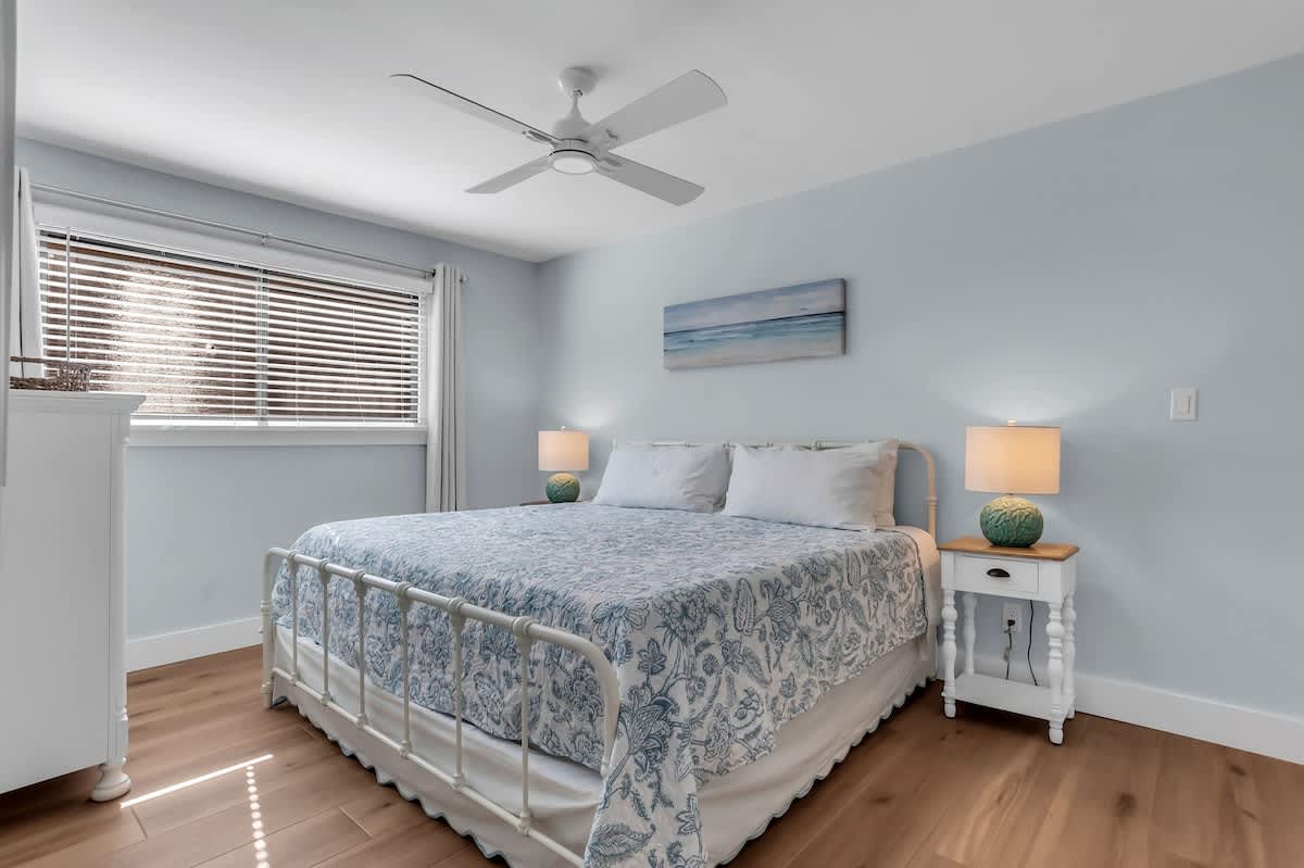 A welcoming guest bedroom designed with soft coastal tones and calming natural light. The comfortable bed is dressed in airy blue-and-white linens, complemented by matching nightstands and warm bedside lighting.