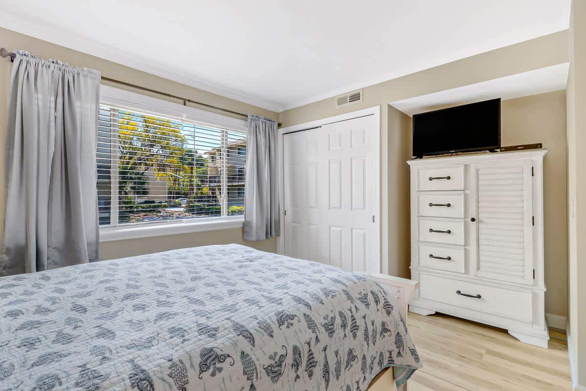 Unwind in your serene bedroom featuring a comfortable queen bed, charming white dresser with TV, and peaceful garden views through your private window.