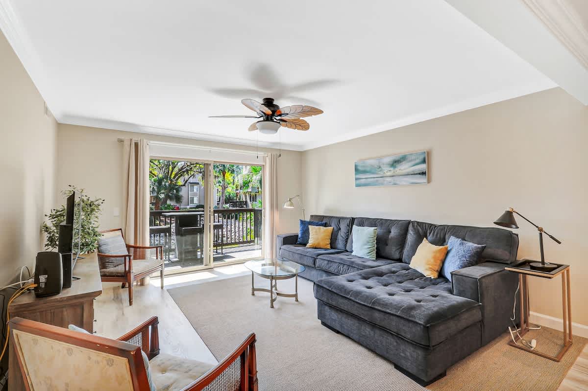 Unwind in your bright living space featuring a plush sectional sofa and tropical ceiling fan, with French doors opening to your private balcony.
