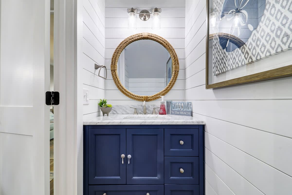 Step into your coastal-themed bathroom featuring a striking navy vanity, rope-framed mirror, and charming shiplap walls that create a serene nautical retreat.