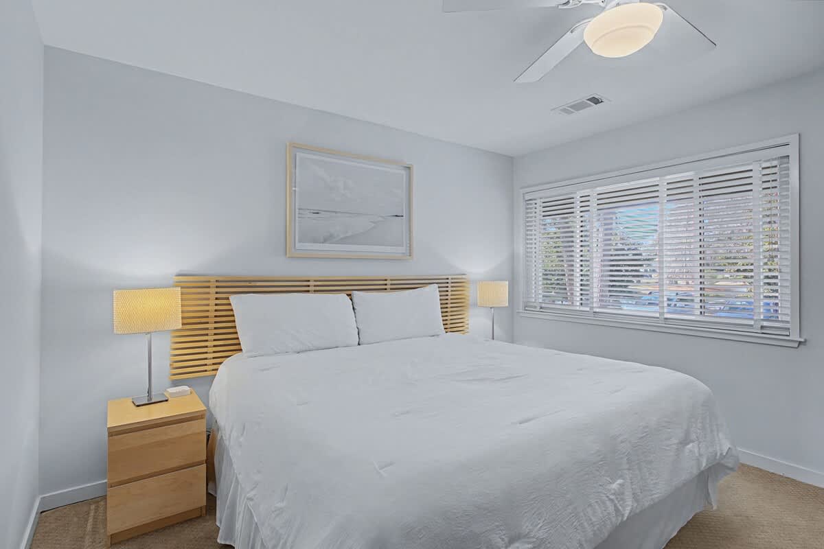 Rest peacefully in your modern bedroom featuring crisp white linens, a warm wood headboard, and natural light filtering through window blinds.