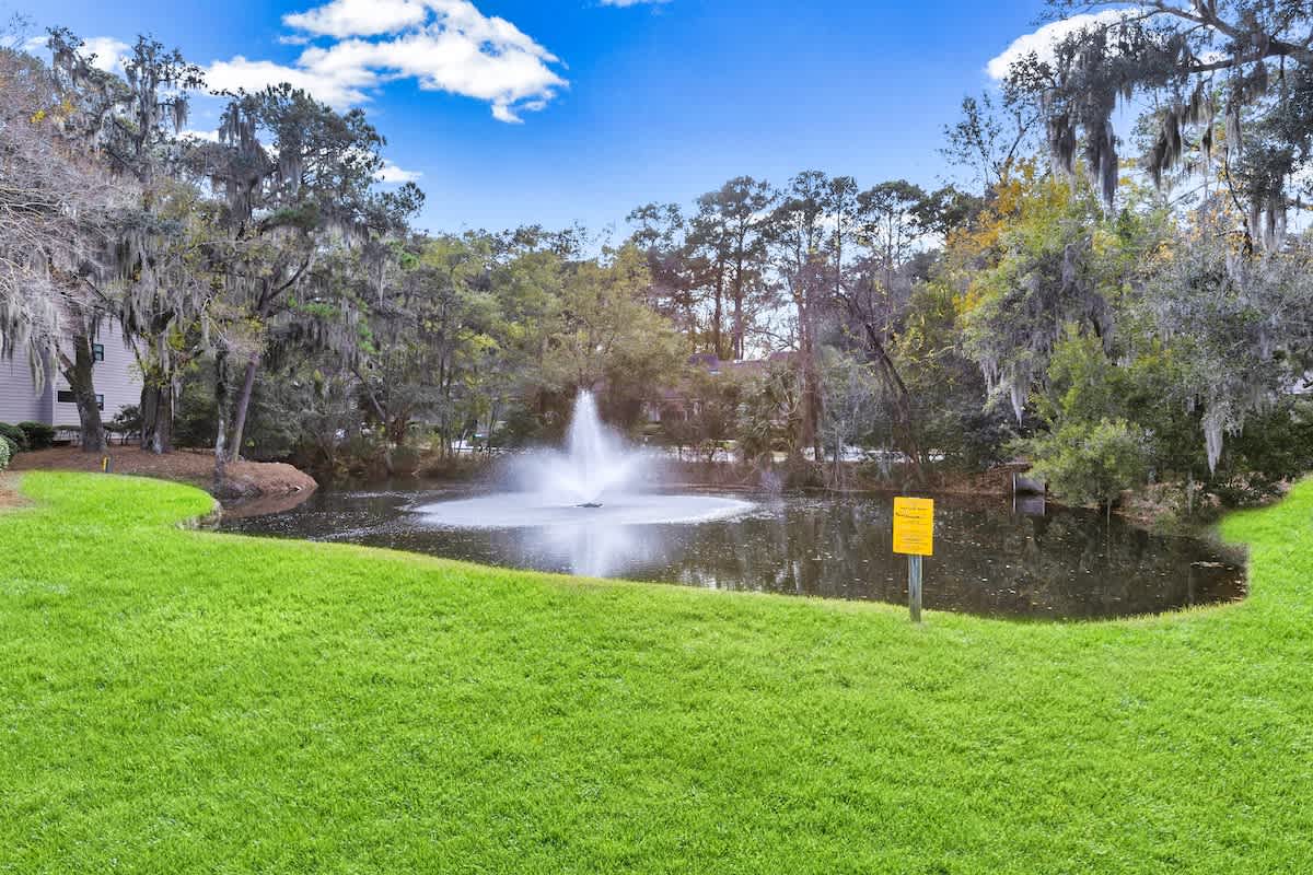 Peaceful pond with fountain surrounded by lush green space and mature trees draped with Spanish moss.