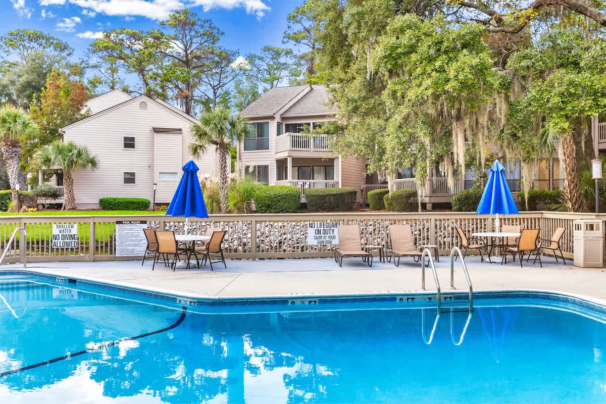Sparkling community pool with lounge chairs and umbrellas, surrounded by lush tropical landscaping and residential buildings.