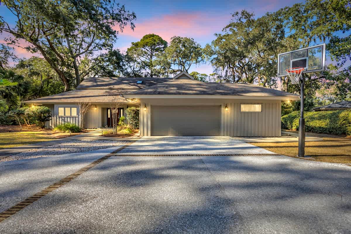 Modern home nestled among mature trees with basketball court and spacious driveway under colorful twilight sky.