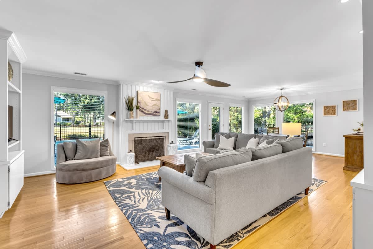 Sink into plush gray seating by your cozy fireplace, where hardwood floors gleam and natural light streams through windows overlooking the pool area.