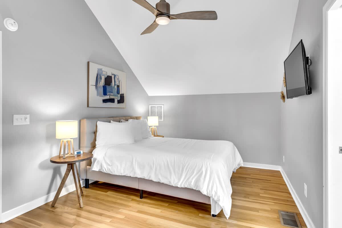 Drift off in your cozy attic bedroom, featuring soft gray walls, warm wood floors, and modern touches like the ceiling fan overhead.