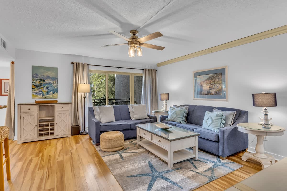 Relax in your bright, coastal-inspired living space with plush blue seating, nautical décor, and peaceful garden views through the large windows.