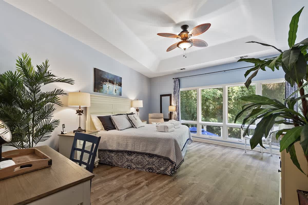 Retreat to your serene bedroom featuring lush tropical plants, a cozy ceiling fan, and bright windows overlooking peaceful greenery.