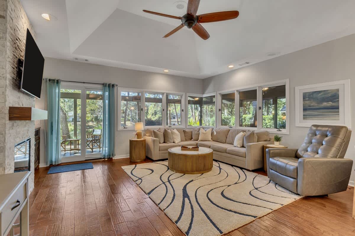 Unwind in this beautifully appointed living room with soaring ceilings, cozy fireplace, and wraparound windows showcasing lush greenery.
