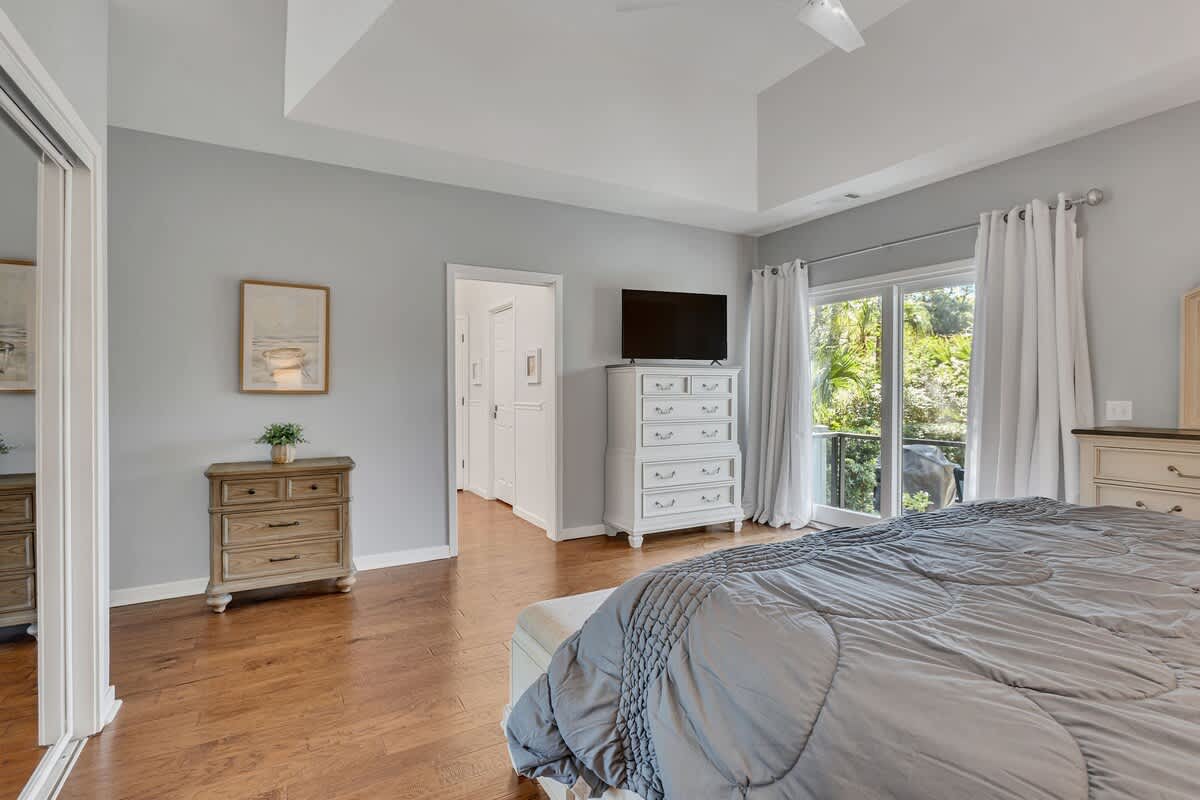 Relax in your spacious bedroom featuring elegant gray walls, warm hardwood floors, and large windows overlooking lush greenery.