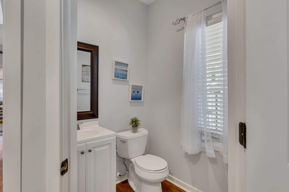Your private powder room features crisp white finishes and coastal artwork, creating a peaceful retreat with natural light streaming through shuttered windows.