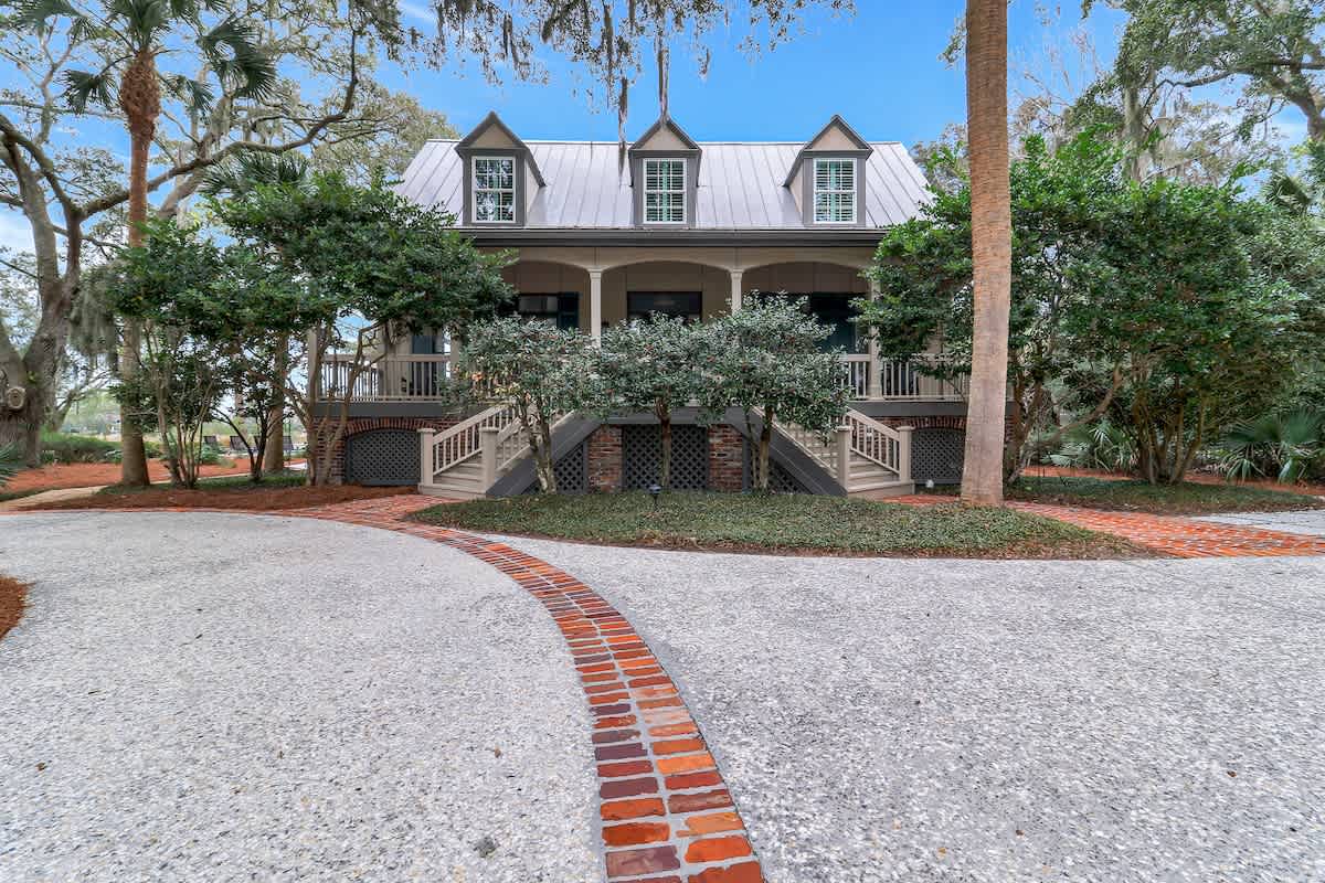 Welcome to this classic Lowcountry home, featuring a raised front porch, charming dormer windows, and a graceful brick-accented driveway framed by mature trees and lush coastal landscaping.