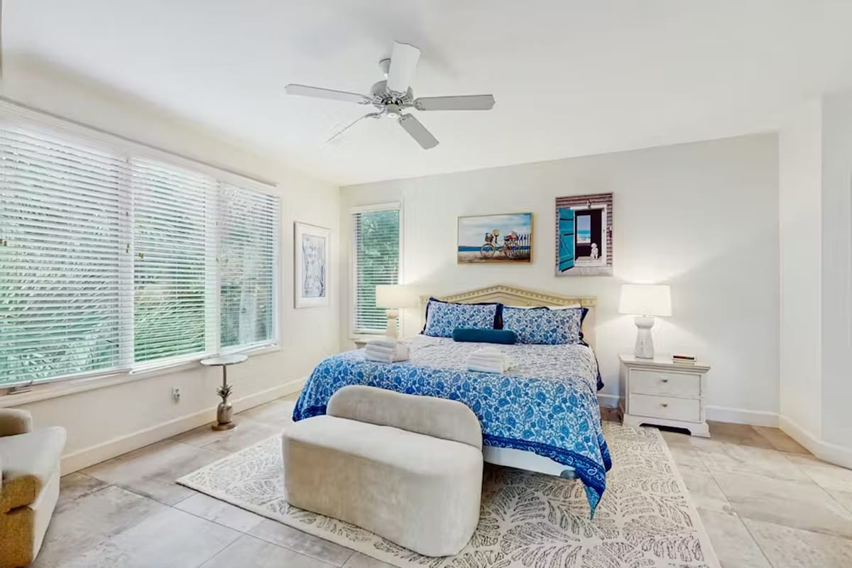 Unwind in your serene bedroom featuring ocean-inspired blue linens, gentle ceiling fan breezes, and peaceful garden views through large windows.
