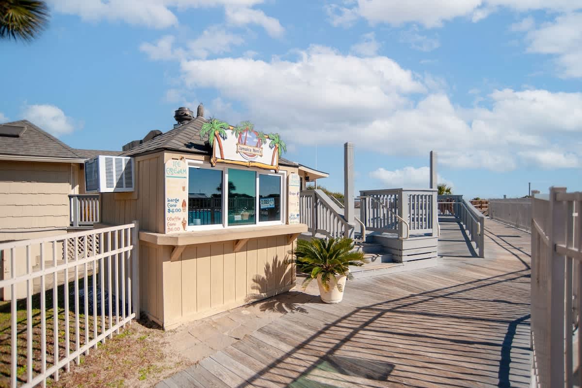 Beachside boardwalk leading to rental shop and beach access, surrounded by coastal homes and palm trees under sunny skies.