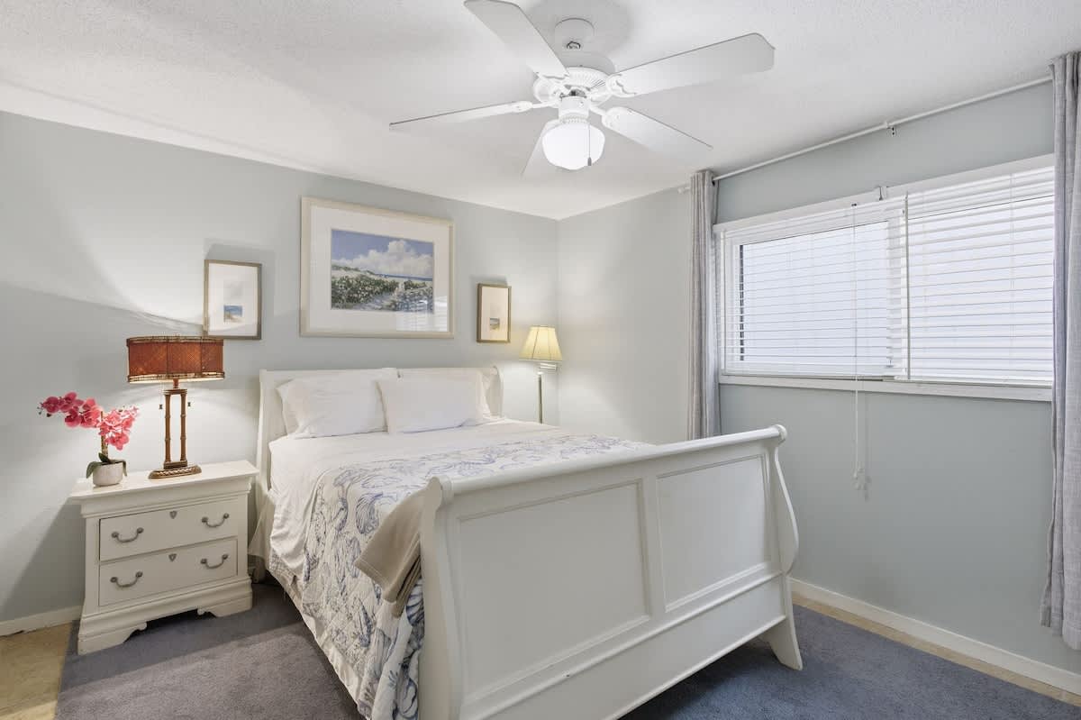 Rest peacefully in your serene bedroom featuring a comfortable white sleigh bed, ceiling fan for perfect temperature, and soft natural light filtering through blinds.