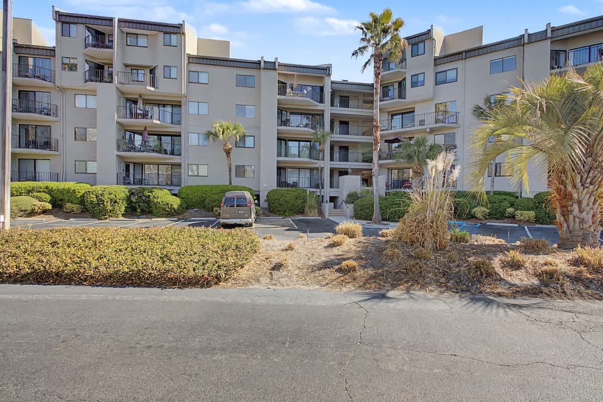 Modern apartment complex featuring multiple levels with private balconies, surrounded by tropical palm trees and landscaped grounds with convenient parking access.