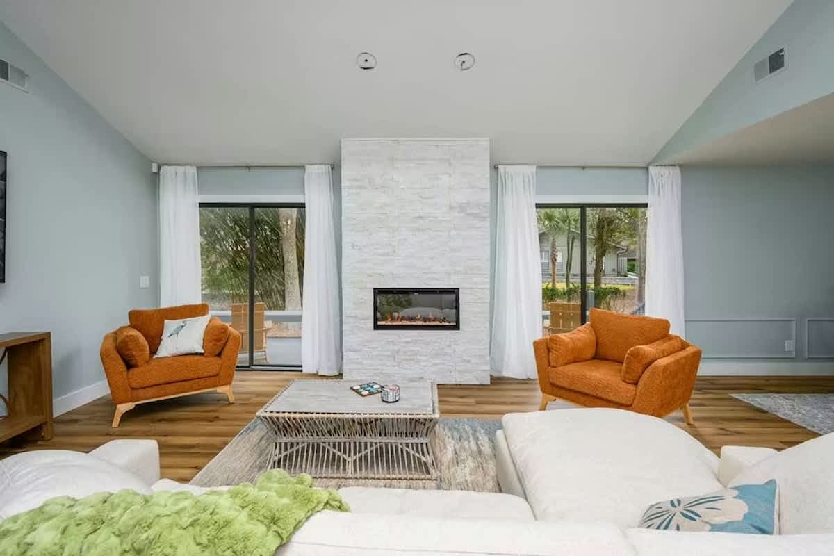 Relax in your stylish living room with modern fireplace, cozy orange armchairs, and peaceful garden views through floor-to-ceiling windows.