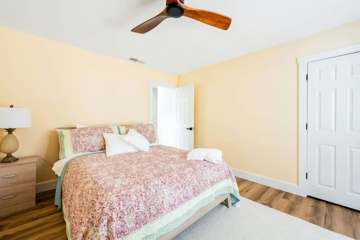 Your cozy bedroom features a comfortable bed with floral linens, warm yellow walls, and a ceiling fan for comfort.
