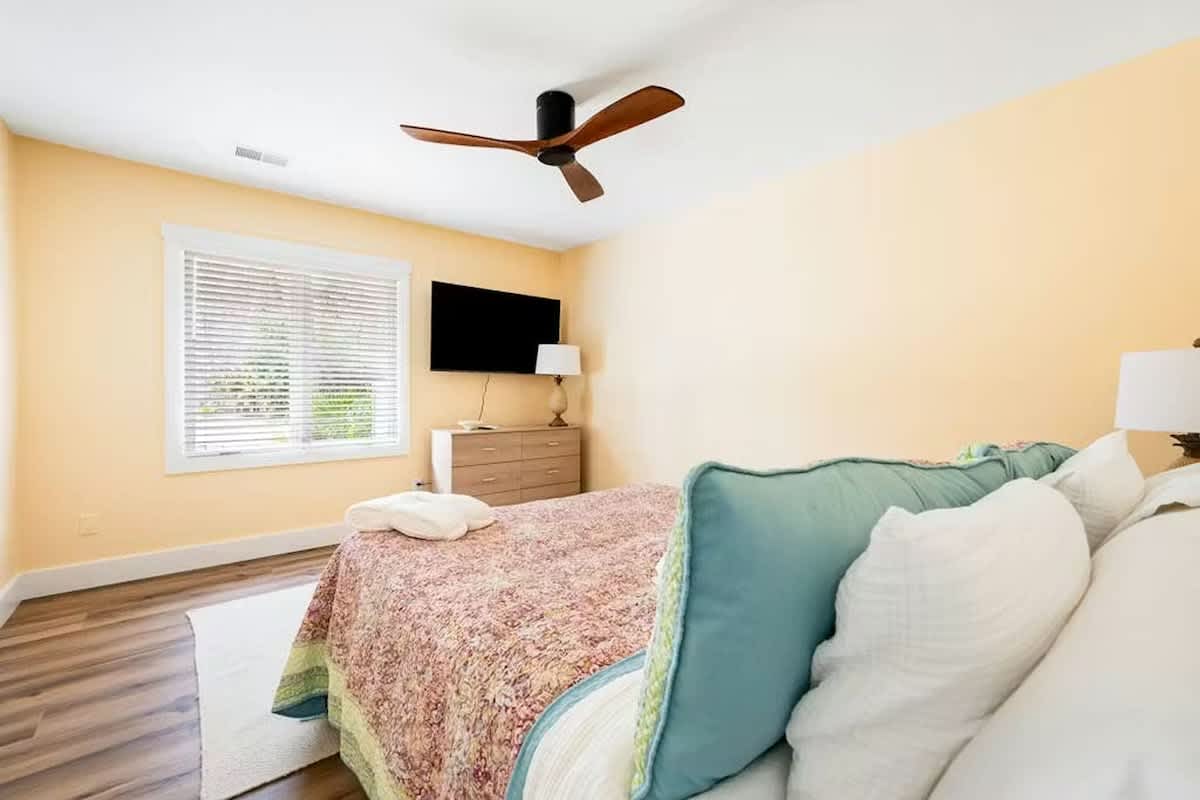 Rest peacefully in your bright, airy bedroom featuring cozy linens, ceiling fan comfort, and tranquil garden views through blinds.