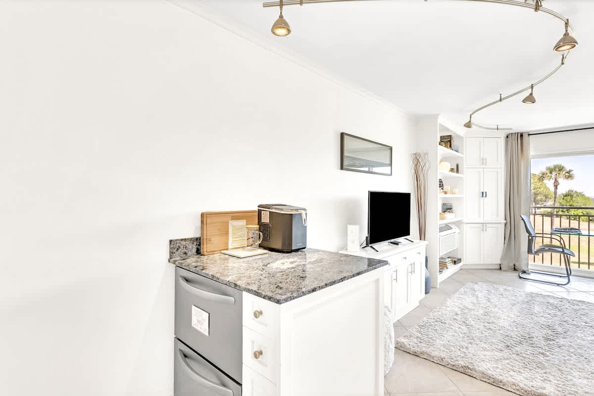 Your bright kitchenette with granite counters and modern appliances opens to a cozy living space with balcony views of palm trees.