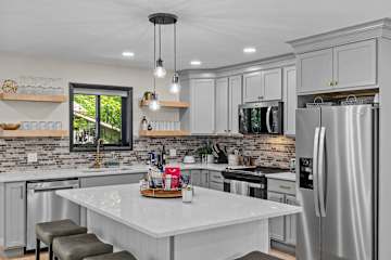 Gather around the white quartz island and cook for real — stainless appliances, open wood shelving with glassware already staged, and a pendant cluster above that makes the kitchen feel less like effort.