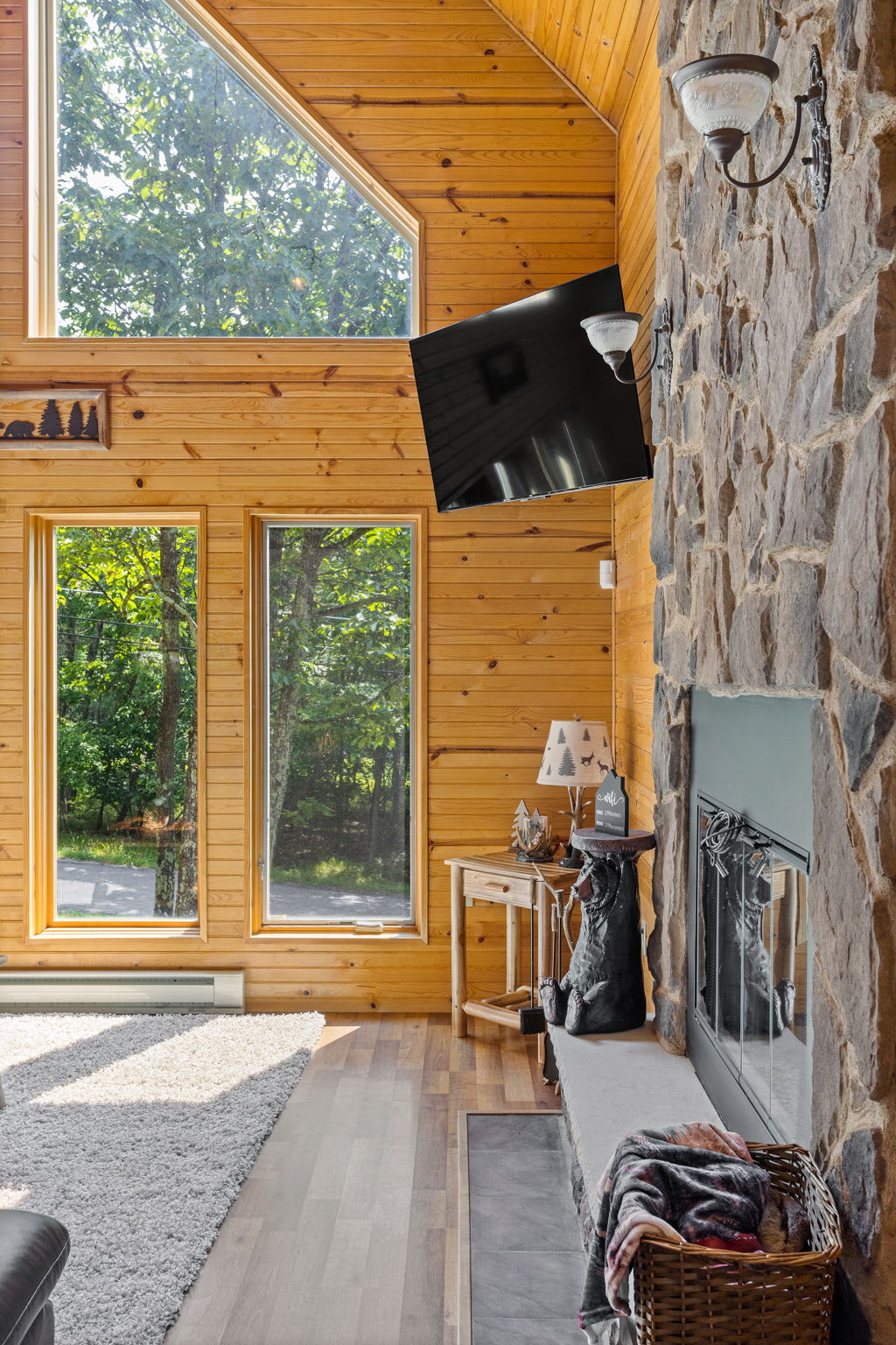 The rich stone fireplace and natural wood interior create a cozy and inviting space, complemented by floor-to-ceiling windows that bring in the beauty of the outdoors.