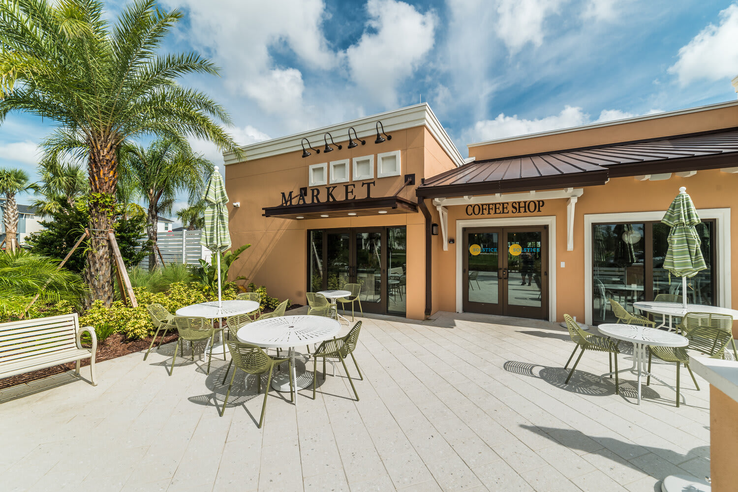 Outdoor seating area with tables and umbrellas in front of a market and coffee shop under a bright sky.
