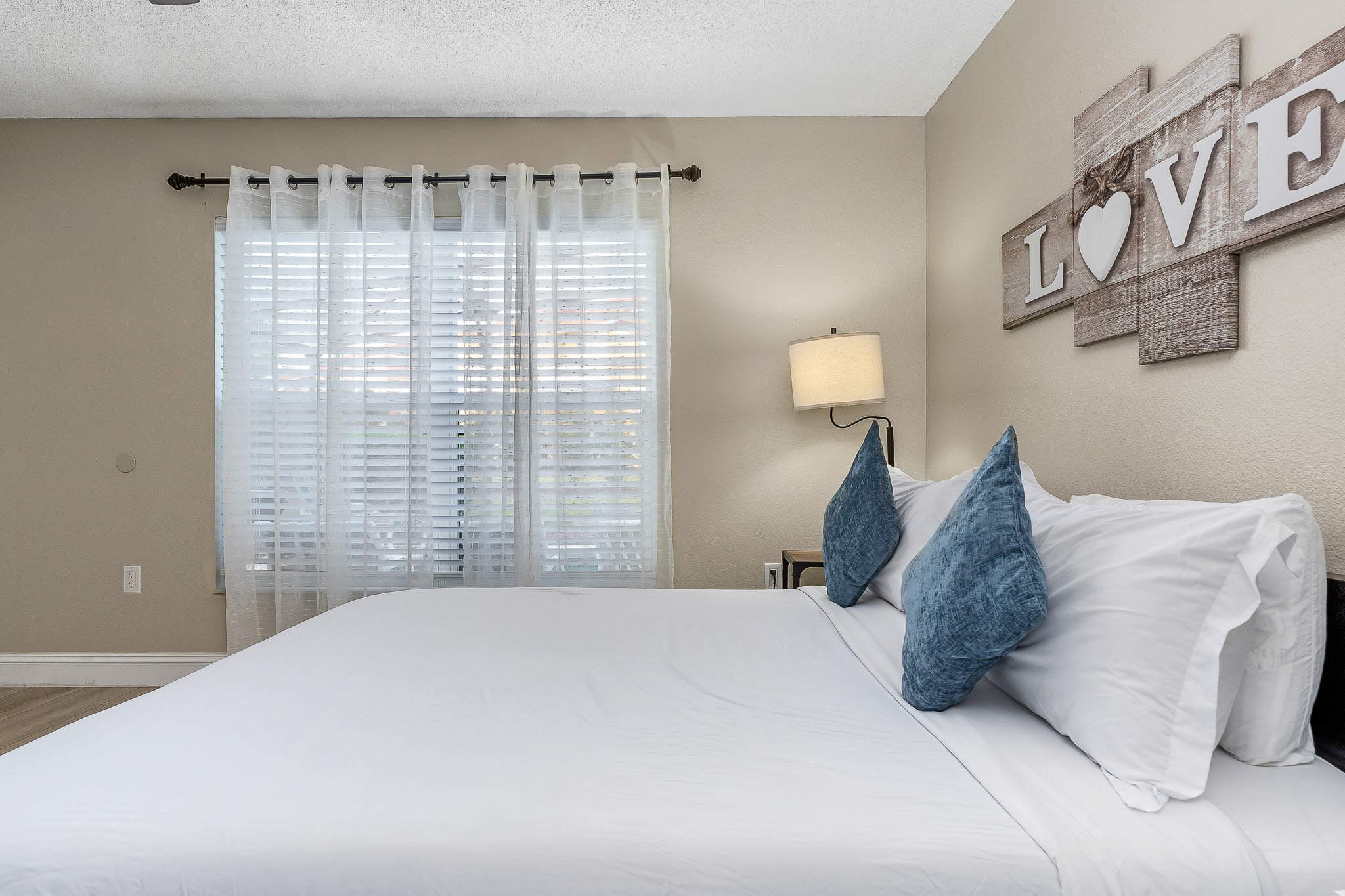 Serene bedroom w/ soft linens, layered pillows, and filtered daylight through sheer curtains.