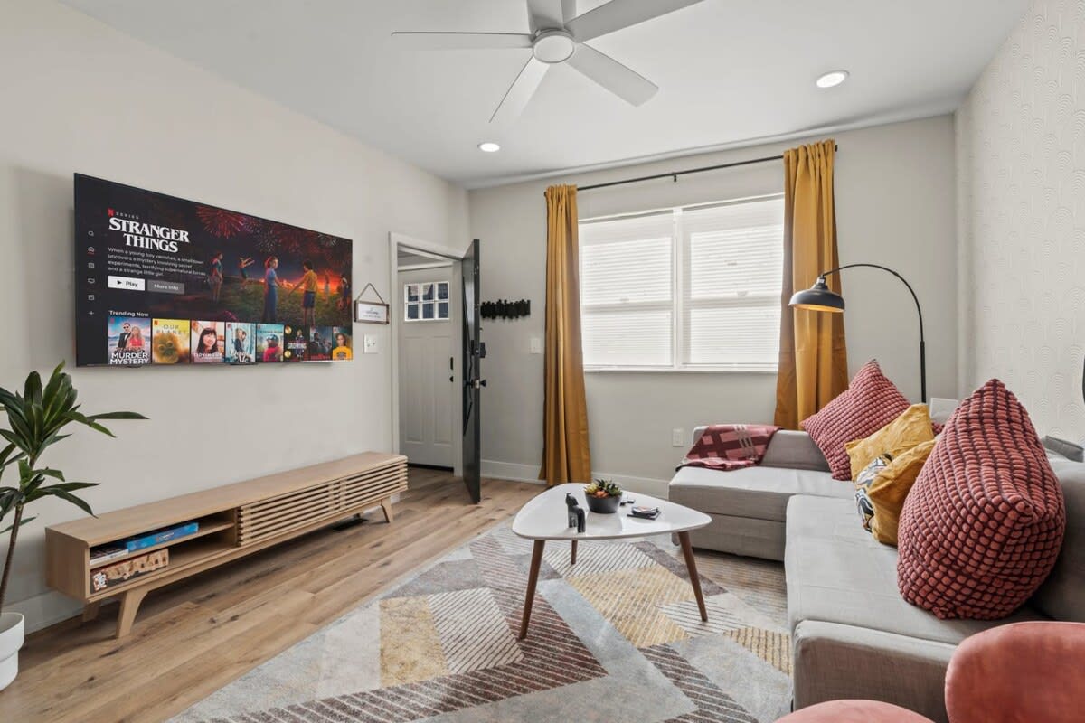 The mustard yellow curtains, floor lamp, and potted plants enhance the cozy ambiance. The TV is wall-mounted, and the media console below offers additional storage and a streamlined look.