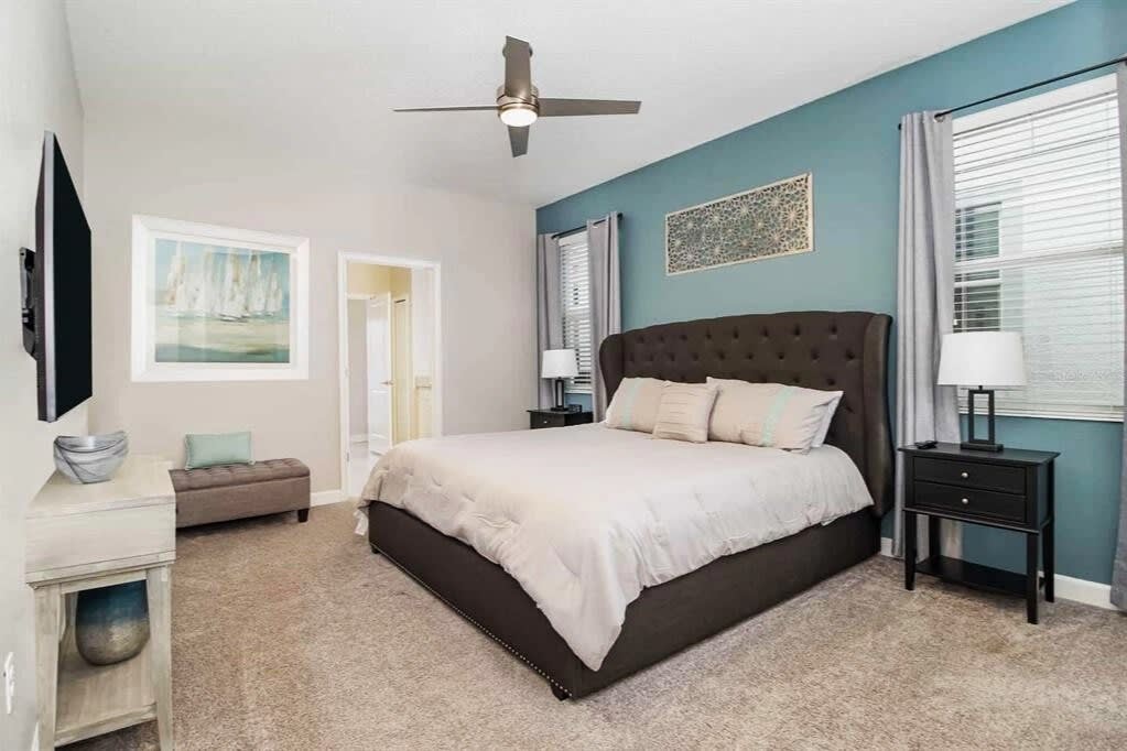 Serene king bedroom with a tufted headboard, soft blue accent wall, coastal artwork, and bright windows framed by light gray curtains.