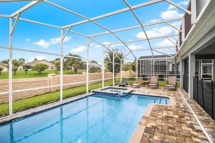 Private screened pool with an attached hot tub, loungers, and a spacious stone-paver deck overlooking sunny green surroundings.