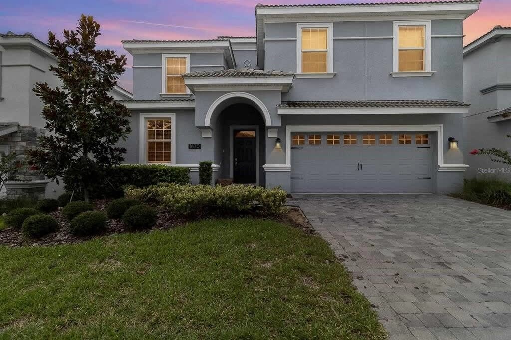 Villa entrance with a graceful arched doorway, warm-lit windows, manicured landscaping, and a spacious paver driveway beneath a colorful twilight sky.